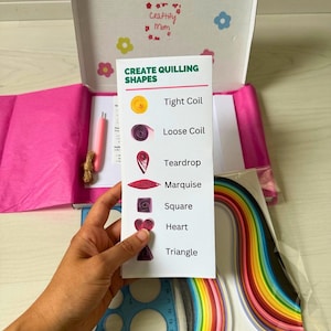May include: A quilling craft kit with a box decorated with flowers and the text "Craftify Mom." The kit contains a guide to create shapes such as tight coils and teardrops, along with colourful paper strips and a pink pen.