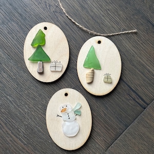 May include: Three wooden Christmas ornaments with sea glass designs. Two ornaments feature green glass trees and small gift boxes. The third ornament has a snowman made of white and clear glass with a carrot nose and green glass scarf.
