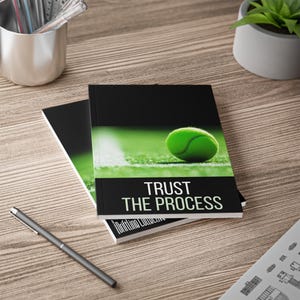 May include: A notebook with a black cover featuring a green tennis ball on a court and the white text "TRUST THE PROCESS" printed on the front. The notebook is positioned on a wood-grain surface next to a pen and a metal cup containing pens.
