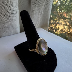 May include: A gold-toned ring featuring an oval-shaped iridescent shell cabochon center stone. The metal setting displays intricate filigree detailing around the perimeter of the stone, with a smooth, thin band extending from the decorative mounting.