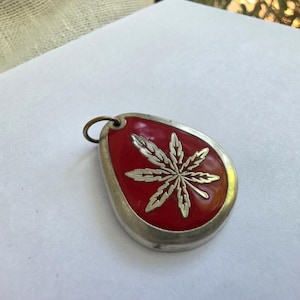 May include: A teardrop-shaped pendant featuring a metallic silver border and a vibrant red enamel center. The center displays a raised, detailed silver leaf design with multiple pointed segments. A small circular metal loop is attached at the top.