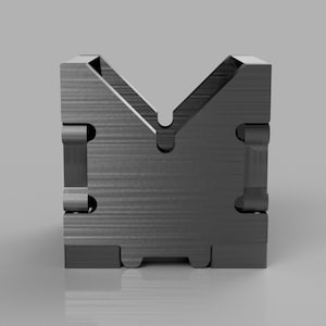 Puede incluir: Un bloque en V de metal negro con una superficie lisa y texturizada. El bloque tiene una ranura en forma de U en el centro y está diseñado para sujetar y sostener objetos.