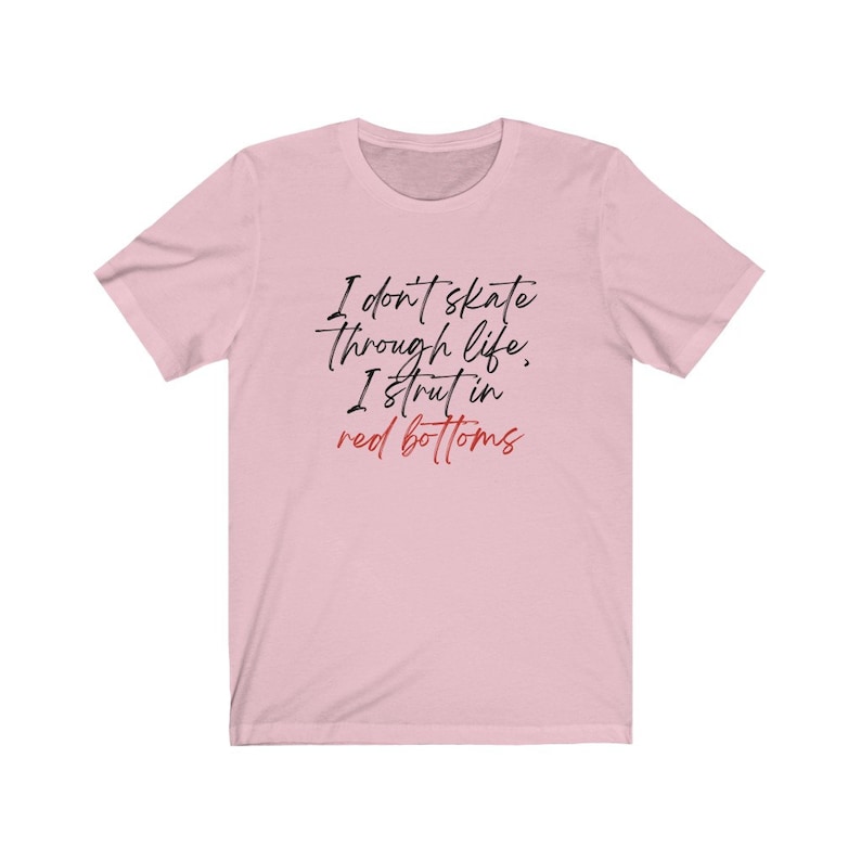 以下が含まれることがあります： ピンクのTシャツに「I don't skate through life, I strut in red bottoms」という黒い文字と赤い文字で書かれたテキストがプリントされています。