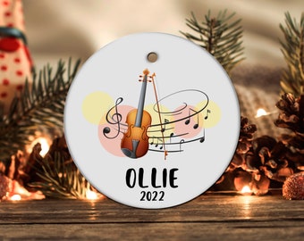 May include: A white round Christmas ornament with a brown violin, musical notes, and the text "OLLIE 2022".