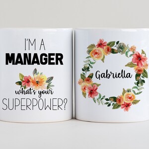 May include: Two white coffee mugs with black text and floral designs. One mug says "I'm a Manager, what's your superpower?" and the other mug has a floral wreath with the name "Gabriella" in the centre.