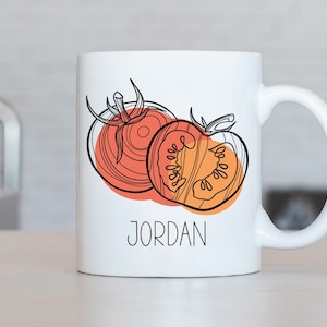 May include: White ceramic mug with a black line drawing of two tomato halves. The tomato on the left is whole, and the tomato on the right is cut in half. The mug has the name "JORDAN" printed in black on the front.