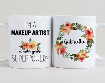 Può includere: Due tazze da caffè bianche con testo nero e disegni floreali. Una tazza dice "Sono una truccatrice, qual è il tuo superpotere?" e l'altra tazza ha una ghirlanda di fiori con il nome "Gabriella" al centro.