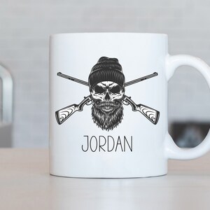 Puede incluir: Taza de cerámica blanca con una ilustración en blanco y negro de una calavera con un gorro y barba, sosteniendo dos rifles cruzados. El texto "JORDAN" está impreso debajo de la ilustración.