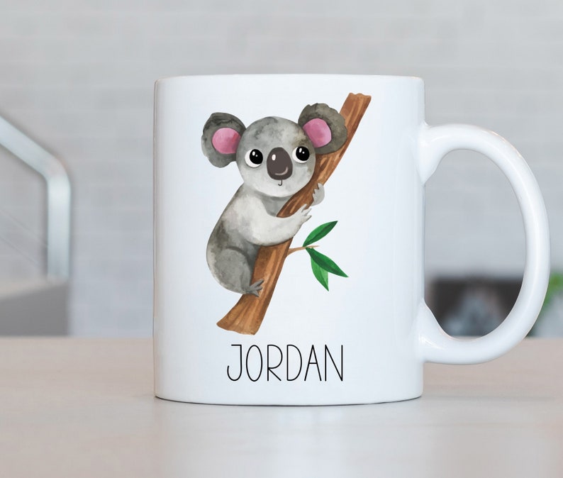 May include: White ceramic mug with a watercolor illustration of a koala bear holding onto a tree branch. The mug has the name "JORDAN" printed on the bottom.