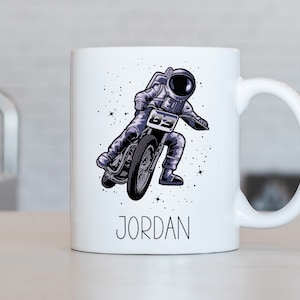May include: White ceramic mug with a graphic of an astronaut riding a motorcycle in space. The mug has a black handle and the name "JORDAN" printed on the bottom.