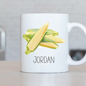 Op de afbeelding: Witte keramische mok met een afbeelding van maïs en de naam "JORDAN" in het zwart gedrukt.
