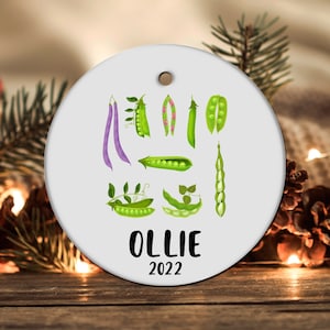 May include: White round Christmas ornament with a green pea pod design and the name "OLLIE" and the year "2022" printed in black.