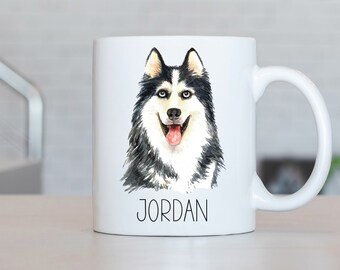 May include: White ceramic mug featuring a watercolor illustration of a Siberian Husky dog with blue eyes and a pink tongue. The name "JORDAN" is printed in black below the dog's image. The mug has a curved handle.