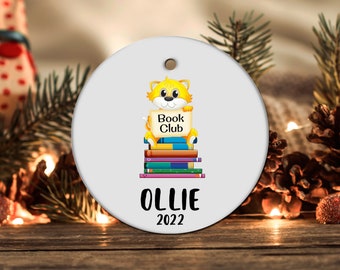 May include: A white round Christmas ornament with a yellow cat sitting on a stack of colorful books. The cat is holding a sign that says "Book Club". The ornament also has the name "OLLIE" and the year "2022" printed on it.