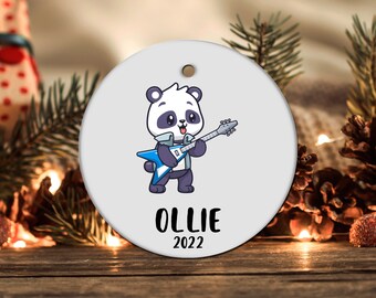 May include: A white round Christmas ornament with a cartoon panda playing a blue electric guitar. The ornament has the name "OLLIE" and the year "2022" printed on it.