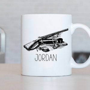 Może przedstawiać: Biały kubek ceramiczny z czarno-białym rysunkiem karabinu, kompasu i karafki. Kubek ma nadrukowane imię "JORDAN" z przodu.