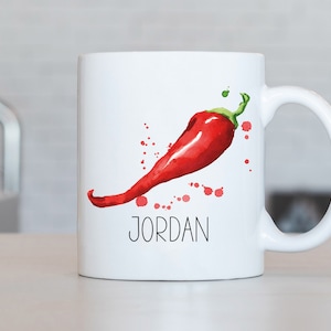 Può includere: Tazza in ceramica bianca con un'illustrazione di peperoncino rosso e schizzi di acquerello rosso. La tazza ha un manico nero e il nome "JORDAN" stampato in nero sotto il peperoncino.
