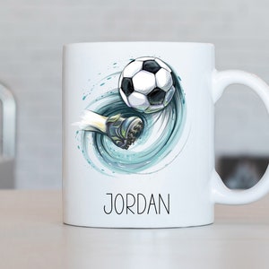 Może przedstawiać: Biały kubek ceramiczny z grafiką piłkarza kopiącego piłkę. Grafika jest w stylu akwarelowym w kolorze niebieskim i zielonym. Kubek ma nadrukowane imię "JORDAN" na dole.