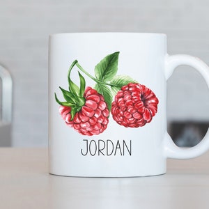 Peut inclure: Une tasse en céramique blanche avec une illustration aquarelle de deux framboises rouges et de feuilles vertes. La tasse porte le nom "JORDAN" imprimé sur le devant.