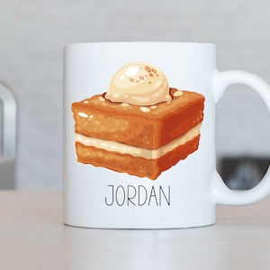 May include: White ceramic mug with a cartoon illustration of a slice of cake with frosting and the name "JORDAN" printed below.