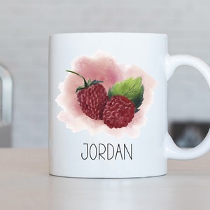 Peut inclure: Une tasse en céramique blanche avec un motif aquarelle de deux framboises rouges et une feuille verte. La tasse porte le nom "JORDAN" imprimé sur le devant.