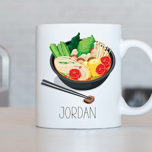 Puede incluir: Taza de cerámica blanca con una ilustración colorida de un tazón de fideos ramen. La taza tiene el nombre "JORDAN" impreso en la parte delantera.