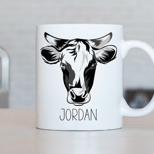 May include: White ceramic mug with a black and white illustration of a cow's head. The mug has a black handle and the name "JORDAN" printed in black below the illustration.