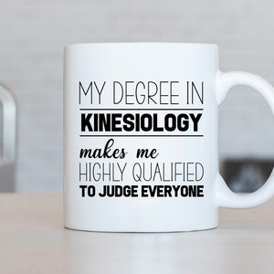 May include: White ceramic coffee mug with black text that reads "My degree in Kinesiology makes me highly qualified to judge everyone."