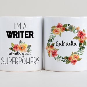 May include: Two white coffee mugs. One mug has the text "I'm a Writer what's your superpower?" with a small floral design. The other mug has a floral wreath with the name "Gabriella" in the center.