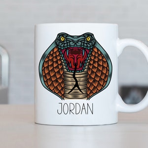 Op de afbeelding: Witte keramische mok met een kleurrijke illustratie van een kobra slang met zijn bek open en zichtbare tanden. De tekst "JORDAN" is onder de afbeelding gedrukt.