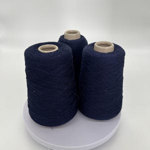 May include: Three navy blue yarn cones are displayed on a white surface. The yarn is tightly wound around cardboard cores. The cones are arranged in a triangular formation, showcasing the texture and color of the yarn. The background is plain white.