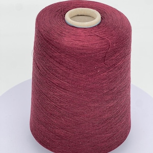 May include: A large spool of burgundy-colored yarn. The yarn is tightly wound around a cardboard core with a light blue and beige rim. The yarn is a solid color and appears to be made of a soft, natural fiber. Ideal for knitting and crochet projects.
