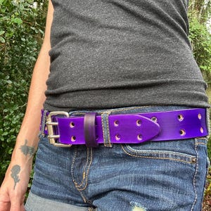 Op de afbeelding: Een paarse leren riem met een zilveren gesp en meerdere gaten. De riem wordt gedragen om de taille van iemand die een spijkerbroek draagt.