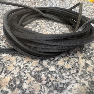 May include: Coiled black leather cord, likely for crafting or repair. The cord is a dark charcoal color and appears to be flexible. The image is taken on a speckled, light-colored surface, providing contrast to the dark cord.