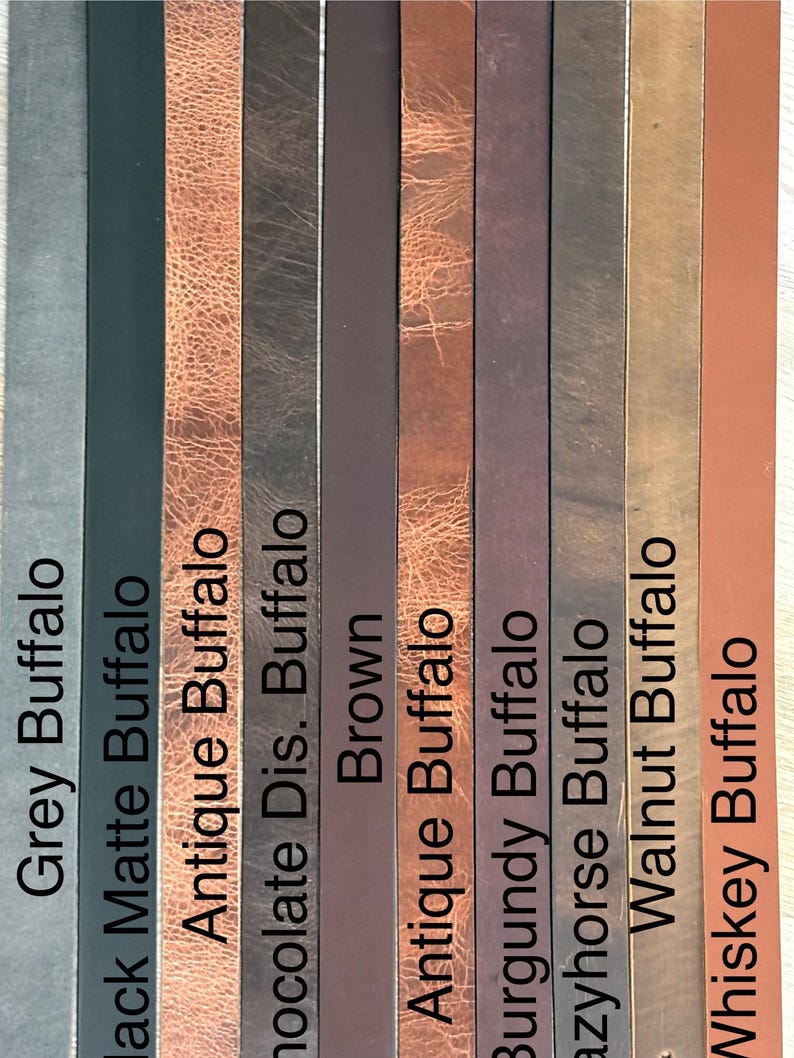 May include: A vertical display of leather strips in various colors, including grey, black, brown, and burgundy. Each strip is labeled with a color name, such as "Grey Buffalo" and "Whiskey Buffalo". The textures vary from matte to distressed.