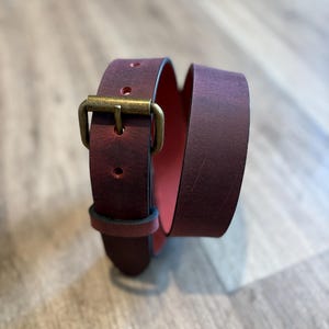 May include: A burgundy leather belt with a brass buckle. The belt is rolled up, showing a pink interior. The leather has a slightly distressed finish. A classic accessory. The belt's length is approximately 100 cm.