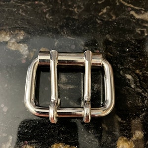 May include: A silver metal buckle with two loops. The buckle is designed for a belt or strap.