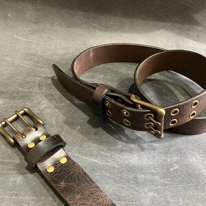 May include: Two brown leather belts with brass buckles and eyelets. One belt has a double row of eyelets and the other has a single row.