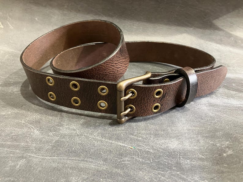 May include: A brown leather belt with a brass buckle and multiple rows of eyelets. The belt is folded in half, showing the buckle and the eyelets.