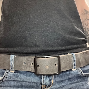 May include: A gray leather belt with a black metal buckle. The belt is worn around the waist and is secured with a buckle.