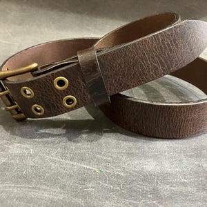 May include: A brown leather belt with a brass buckle and three eyelets. The belt is folded in half, showing the inside of the belt.