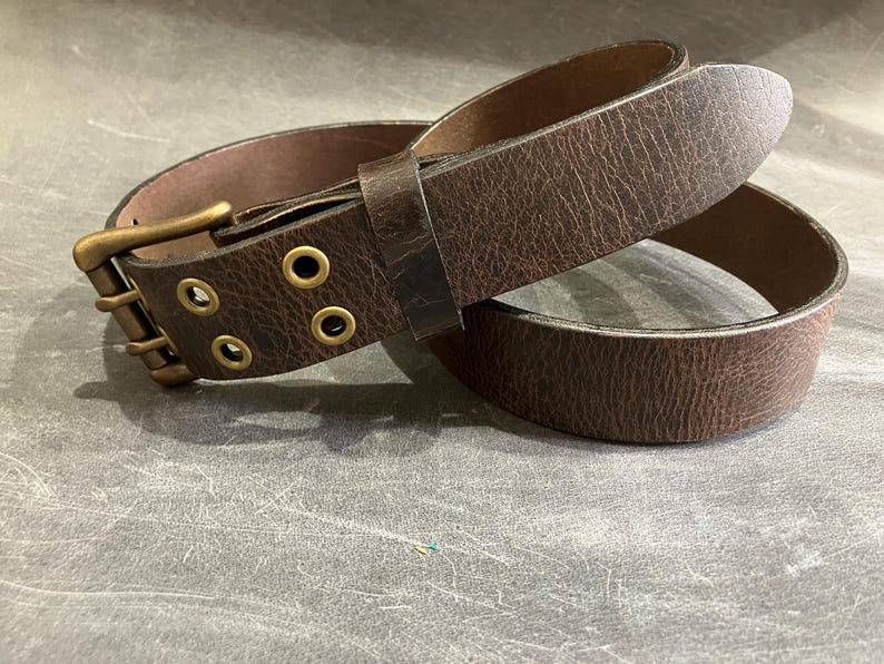 May include: A brown leather belt with a brass buckle and three eyelets. The belt is folded in half, showing the inside of the belt.