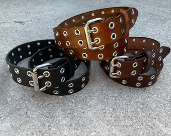 May include: Three leather belts in black, medium brown, and dark brown, each with silver-tone metal grommets and a matching buckle. The belts are arranged in a layered display, showcasing their design and color variations.