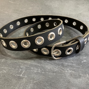 May include: A black leather belt with silver grommets and a silver buckle. The belt is folded in half and the buckle is visible.