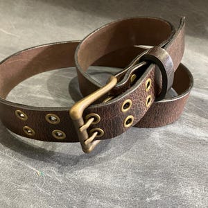 May include: A brown leather belt with a brass buckle and multiple eyelets. The belt is worn and has a distressed look.