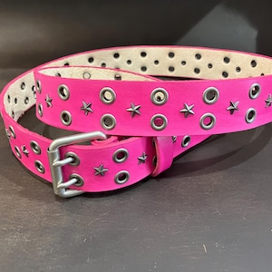 Puede incluir: Un cinturón de cuero rosa con detalles de estrellas plateadas y ojales metálicos. El cinturón es de doble capa y tiene una hebilla.