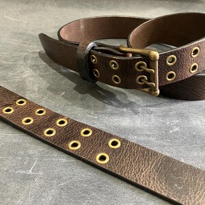 May include: A brown leather belt with a brass buckle and multiple rows of eyelets. The belt is unbuckled and lying flat on a gray surface.