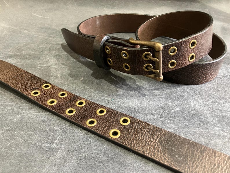 May include: A brown leather belt with a brass buckle and multiple rows of eyelets. The belt is unbuckled and lying flat on a gray surface.