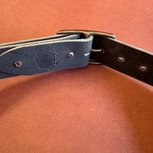 May include: Close-up of a dark blue leather belt with a metal buckle. The belt has a textured surface and is secured with metal rivets. A logo is embossed on the belt near the buckle.