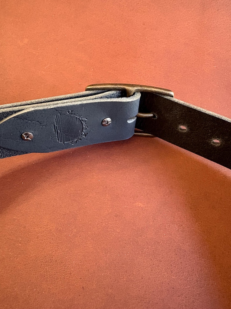 May include: Close-up of a dark blue leather belt with a metal buckle. The belt has a textured surface and is secured with metal rivets. A logo is embossed on the belt near the buckle.